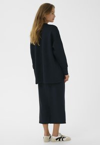 Vrouw met lang haar, gekleed in een zwarte oversized trui, een bijpassende lange rok en witte sneakers met zwarte accenten, staande tegen een effen achtergrond.