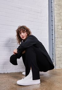 Mladá osoba s kudrnatými vlasy v černém oblečení, která se dřepí uvnitř vedle bílé cihlové zdi, na sobě má bílé vysoké tenisky Calvin Klein.