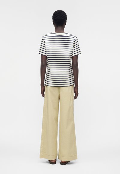 Άτομο που στέκεται με την πλάτη γυρισμένη, φοράει μια λευκή και ναυτικό ριγέ κοντομάνικη μπλούζα και μπεζ παντελόνια με φαρδιά πόδια.