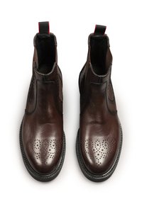Stivali ankle in pelle marrone con perforazioni brogue, dettagli cuciti e una linguetta in tessuto rossa e nera sul retro.