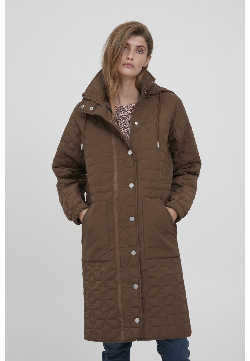 Kvinde iført en lang, brun vatteret frakke med store lommer, sølvfarvede trykknapper og en høj krave, sammen med blå jeans.