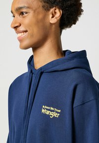 Granatowy sweatshirt z kapturem wykonany z mieszanki bawełny. Na piersi widnieje żółty haftowany napis "W dżinsy wierzymy Wrangler".