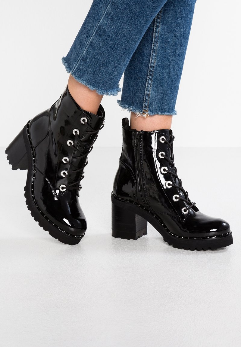 Bottines en cuir verni noir avec talons épais, lacets sur le devant, zips latéraux et œillets argentés, portées avec un jean bleu à ourlets effilochés.