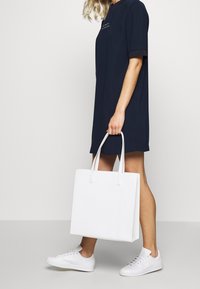 Sac fourre-tout blanc avec logo embossé "TED BAKER LONDON", forme rectangulaire, texture douce et double poignées. Porté avec une robe marine et des baskets.