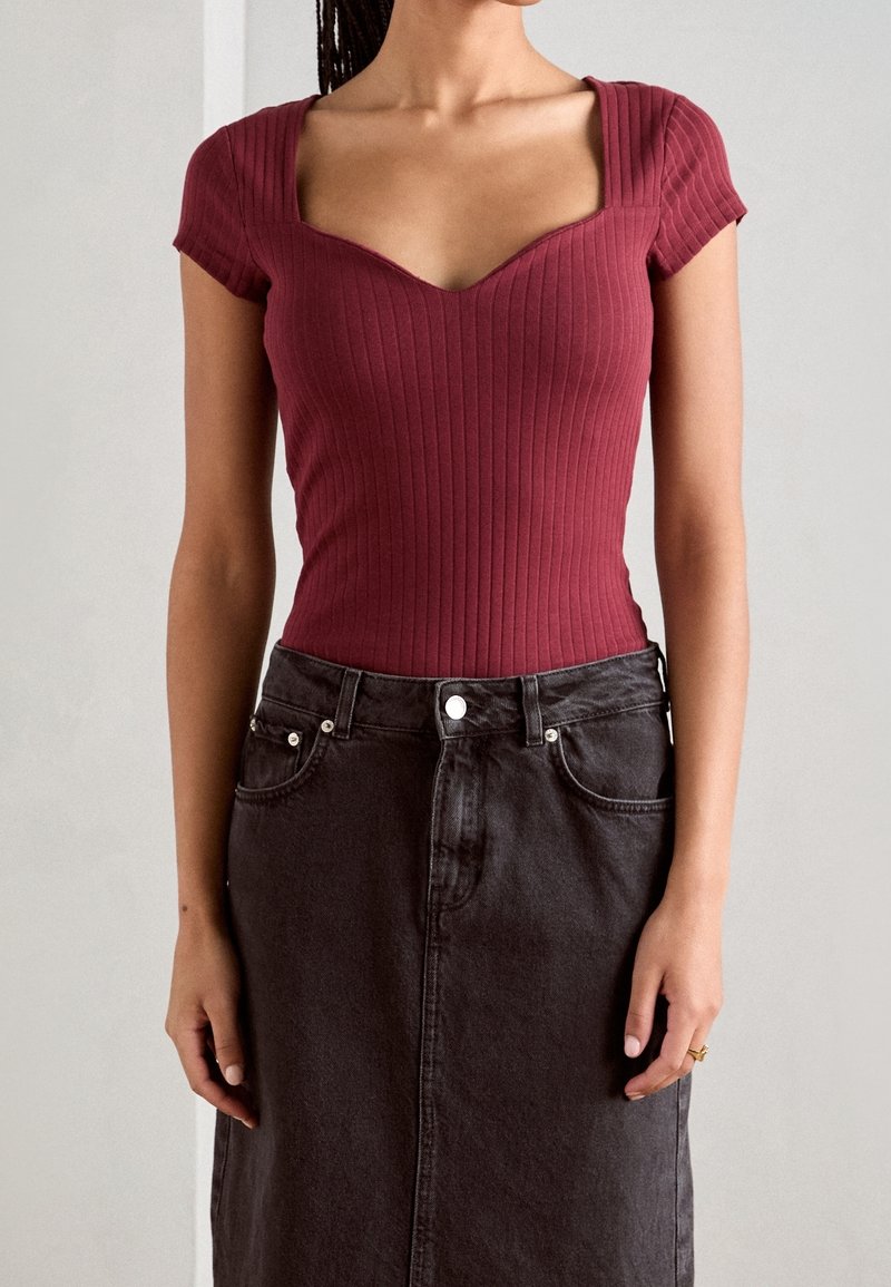 Donna che indossa un top corto a maniche corte con scollatura quadrata di colore marrone bordeaux, infilato in una gonna di jeans nera a vita alta con tasche e passanti per cintura.