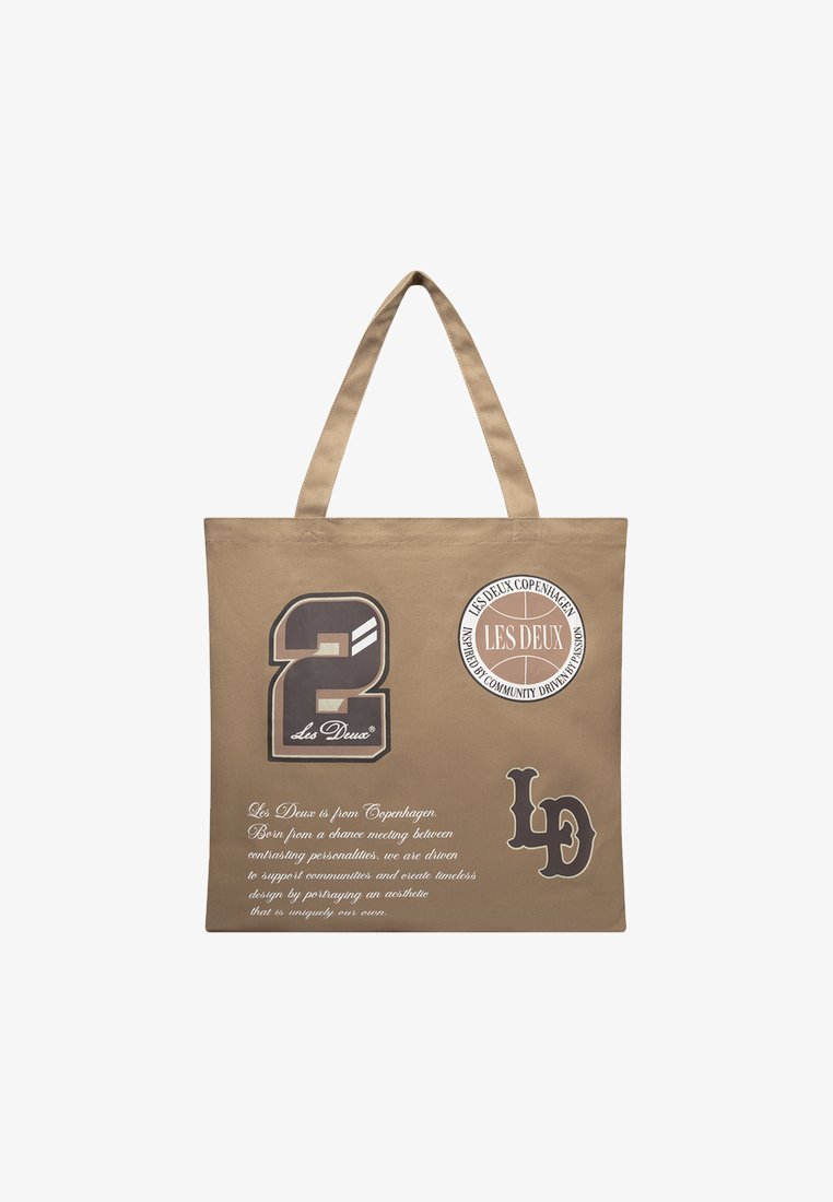Beżowa torba tote z dwoma uchwytami, zdobiona graficznymi nadrukami, w tym cyfrą "2" i "LD" oraz okrągłym logo. Tekst zawiera opis marki.