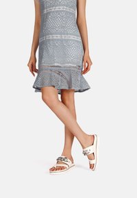 Sandali bianchi con dettagli in catena argentata, dotati di una suola platform. La modella indossa un abito in pizzo azzurro chiaro con orlo a campana.