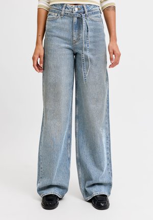 Person trägt hellblaue Jeans mit weitem Bein, einen Stoffgürtel in der Taille gebunden und schwarze Schuhe vor weißem Hintergrund.