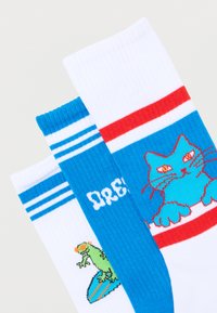 Τρεις πολύχρωμες κάλτσες με μπλε ρίγες, που απεικονίζουν έναν πράσινο κινουμένων σχεδίων σαύρα, τη λέξη "DRE!" και μια μπλε γάτα με κόκκινα μάτια και μουστάκια.