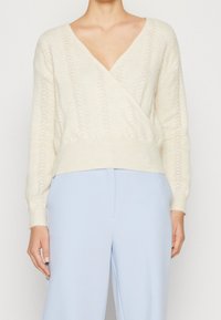 Neulepaita kermaan, jossa syvä V-pääntie, kietaisumalli, teksturoitu kuvio ja leveä resorireuna, yhdistetty vaaleansinisiin housuihin.