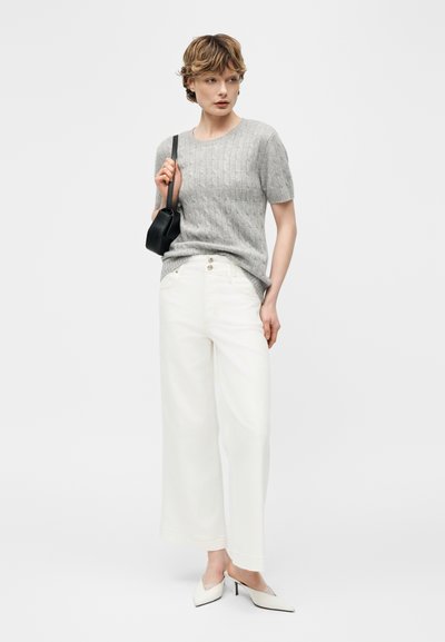 Γυναίκα που φοράει γκρι πλεκτό τοπ με κοντά μανίκια, άσπρο ψηλόμεσο παντελόνι με φαρδιά μπατζάκια, λευκές γόβες και κρατάει μια μικρή μαύρη τσάντα ώμου.