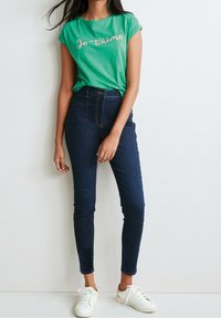 Camiseta verde de manga corta con "Je t’aime" en letras rosas, combinada con jeans skinny azul oscuro y zapatillas blancas.