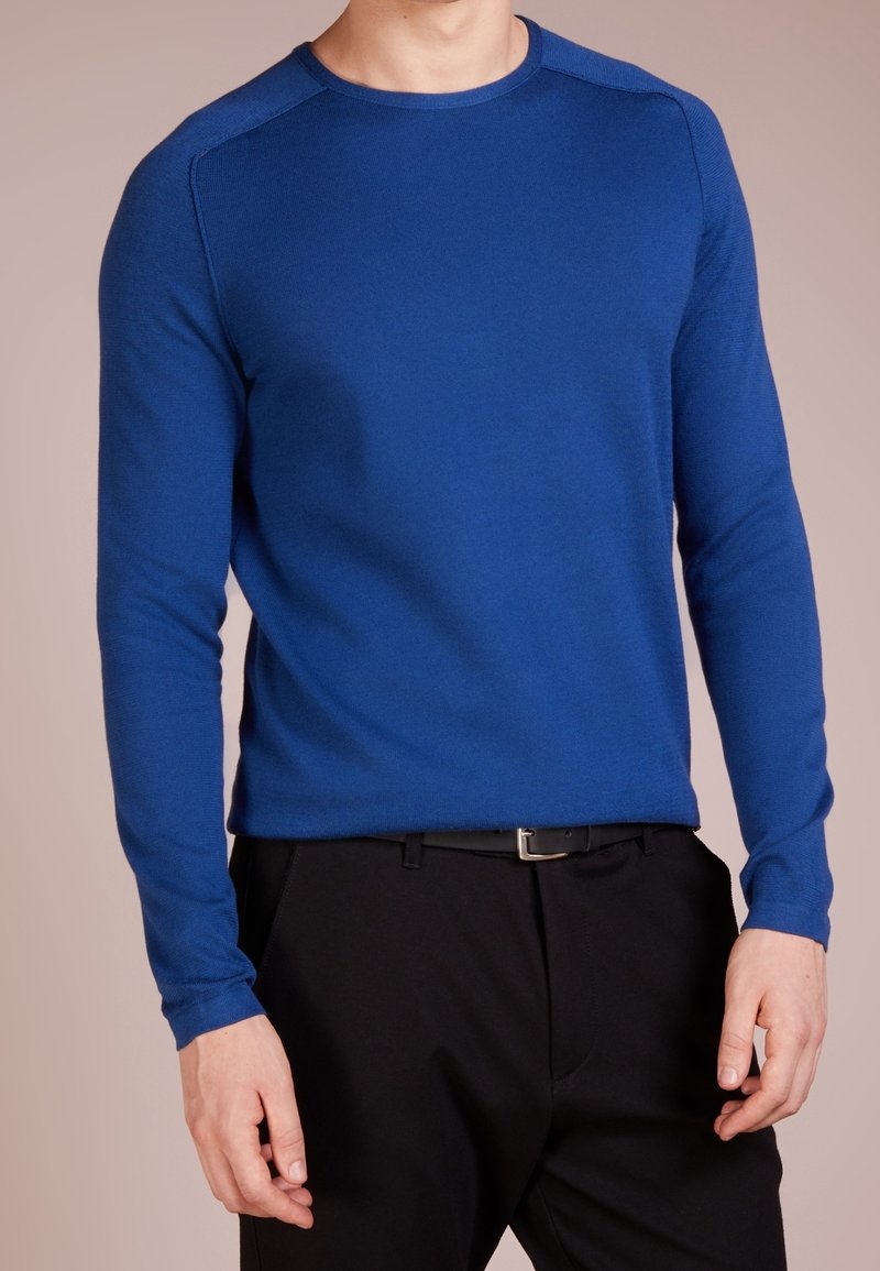 Blue knitted sweater with long sleeves, round neck, and ribbed cuffs; paired with black trousers; smooth texture, fitted design.