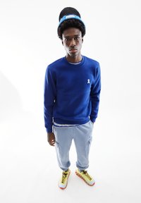 Blauwe crewneck sweatshirt met witte lintdetails en lichtblauwe joggers. Bevat een gelaagde look met een gestreepte inner tee en kleurrijke sneakers.