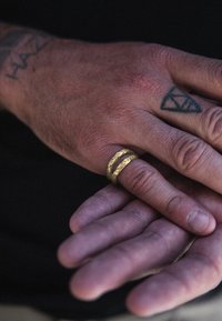 Guldring med en texturerad yta och två staplade band b ärs på en hand med tatueringar, handflatan vänd uppåt, mot en mörk bakgrund.