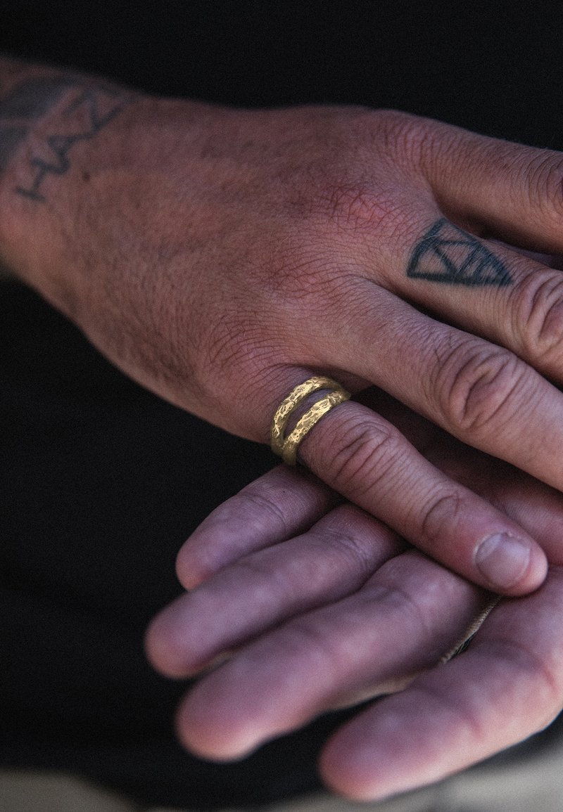 Guldring med en texturerad yta och två staplade band bärs på en hand med tatueringar, handflatan vänd uppåt, mot en mörk bakgrund.