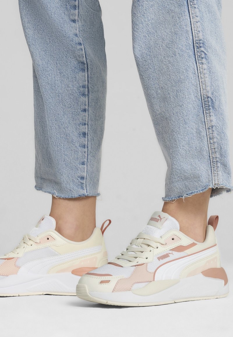 Pieds portant des baskets Puma beige et blanches avec des accents rose clair, associés à un jean en denim bleu clair raccourci.