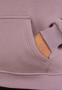 Sweatshirt lilas avec une poche kangourou, des poignets côtelés et une texture douce. Le design présente une coupe ajustée avec un col rond.