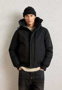 Black puffer jacket with a high collar, front zipper, and side pockets. Worn over a light-colored knit sweater and paired with dark jeans.