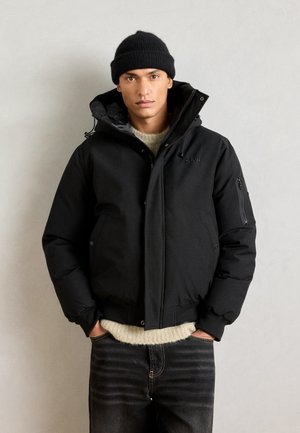 Black puffer jacket with a high collar, front zipper, and side pockets. Worn over a light-colored knit sweater and paired with dark jeans.