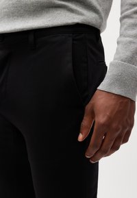 Pantalones negros de corte slim, con bolsillos laterales y una textura suave. Un suéter gris con puños acanalados es parcialmente visible.