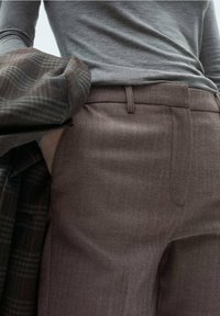 Pantalons marron à motif pied-de-poule avec une texture lisse et des passants de ceinture, associés à un haut gris à manches longues et un blazer à carreaux.