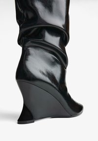 Čierne wedge čižmy vyrobené z lesklej kože, s elegantným siluetom, uvoľneným vrchom a výrazným dizajnom wedge podpätku.