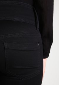 Primer plano de una persona que lleva vaqueros negros con un bolsillo trasero visible y una camiseta de manga larga de canalé negra, contra un fondo neutro.