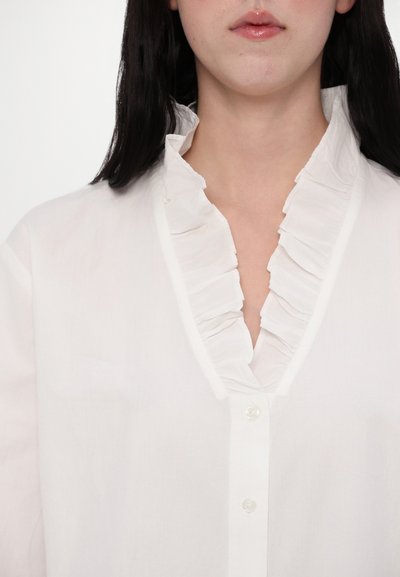 Weiße Bluse mit Rüschkragen, durchgehender Knopfleiste und glatter Textur. Verfügt über zarte Rüschen entlang des Halsausschnitts für zusätzlichen Detail.