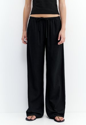 Personne portant un pantalon noir ample à cordon, un top court noir et des sandales, debout devant un fond blanc uni.