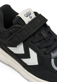 Zapatilla deportiva negra con parte superior de malla, correa de Velcro y detalles en zigzag blancos. Presenta un diseño texturizado y una suela de goma.