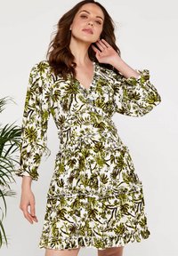 Blumenmusterkleid mit weißem Hintergrund, das grüne und braune Pflanzenmuster, Rüschendetails und lange Ärmel aufweist.