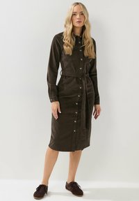 Braunes Cord-Midi-Kleid mit langen Ärmeln, durchgehender Knopfleiste, zwei Brusttaschen und einem Stoffgürtel, kombiniert mit dunklen Sneakers.