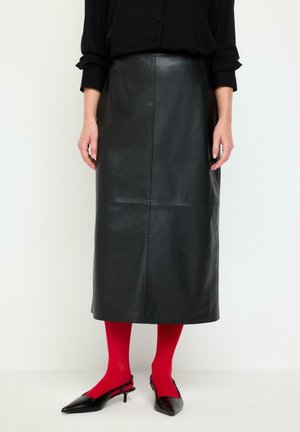Kvinde iført sort lang lædernederdel, sort top, røde strømpebukser og sorte slingback hæle står mod en hvid baggrund.