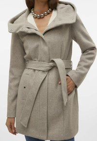 Beigenvärinen villakangastakki hupulla, vyötäröllä vyö, sivutaskut ja nepparikiinnitys. Pehmeä tekstuuri ja mittatilausmainen istuvuus, korostettuna ompelujen yksityiskohdilla.