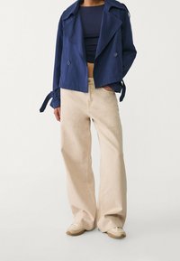 Granatowa krótka kurtka z podwójnymi guzikami i detalami w postaci sznurków, w zestawieniu z beżowymi szerokimi spodniami i kremowymi butami. Gładka faktura materiału.