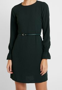 Mörkgrön klänning med långa ärmar, figurnära bodice, utställda manschetter och ett smalt matchande bälte med en metallspänne i midjan.