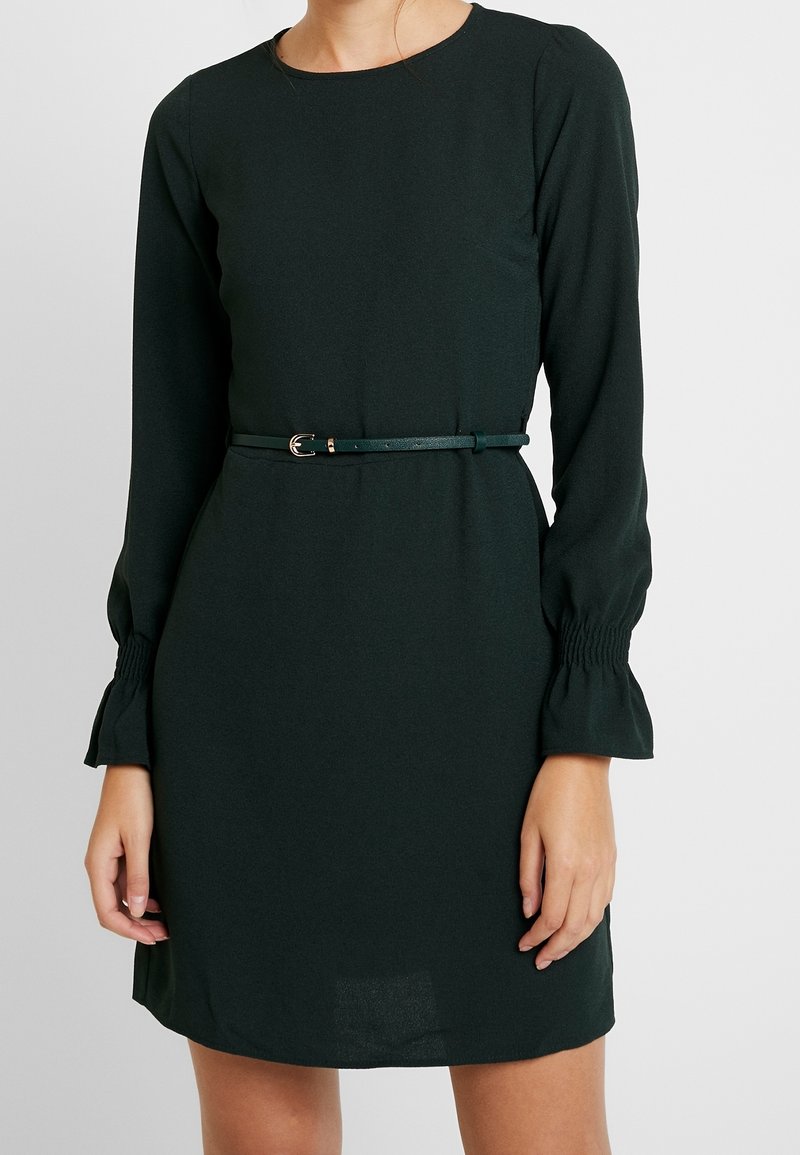 Mörkgrön klänning med långa ärmar, figurnära bodice, utställda manschetter och ett smalt matchande bälte med en metallspänne i midjan.