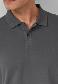 Szara koszulka polo wykonana z teksturowanej tkaniny, z kołnierzykiem, dwoma guzikami i małym logo marki w ciemnoniebieskim kolorze po lewej stronie.