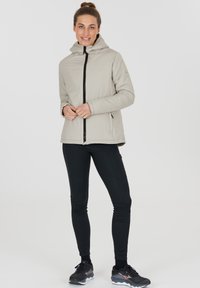 Hellgraue isolierte Jacke mit Kapuze, Frontreißverschluss und schwarzen Akzenten. Kombiniert mit schwarzen, enganliegenden Hosen und Sportschuhen. Glatte Textur.