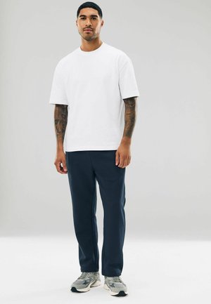 Camiseta blanca de manga corta combinada con pantalones de chándal azul marino. El calzado es de color gris con un patrón texturizado. El modelo posa frente a un fondo sencillo.