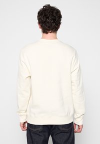 Felpa color crema con maniche lunghe e collo rotondo. Tessuto morbido, polsini e orlo a costine. La vista posteriore mostra un design liscio e minimal.