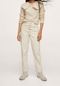 Cardigan en tricot beige avec un large col et un nœud en satin, associé à un pantalon crème taille haute avec un devant à cinq boutons. Baskets légères.