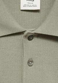 Grå, texturerad polo shirt med ribbad tyg och platt krage. Har två knappar vid halsen. Etiketten visar storleksdetaljer och ursprung.