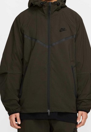 Ciemno oliwkowa kurtka z kapturem, z czarnym zamkiem błyskawicznym, bocznymi kieszeniami i subtelnym czarnym logo Nike na przedniej części klatki piersiowej, noszona przez osobę.