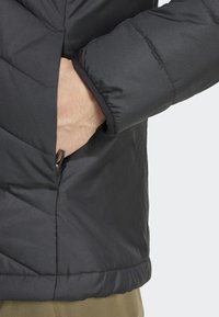 Black padded jacket with a textured finish, featuring a side pocket and a cuffed sleeve. Geometric stitching detail along the body.
