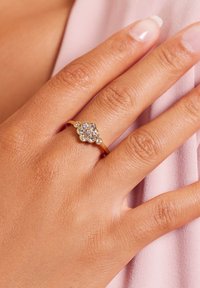 Gouden ring met een cluster van heldere edelstenen, geplaatst in een bloemenschema. Hand zichtbaar tegen lichtroze stof.