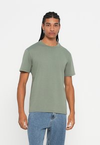 Man with long braided hair wearing a plain sage green short-sleeve T-shirt and light blue jeans, standing against a white background.