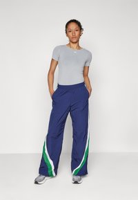 Športový outfit s svetlošedým priliehavým topperom a námorníckymi teplákmi so zelenými a bielymi bočnými pruhmi. Nosené s šedými teniskami.