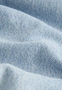 Tissu en denim bleu clair avec un tissage texturé. Matériau doux et fluide avec un relief diagonal visible et une finition lisse.